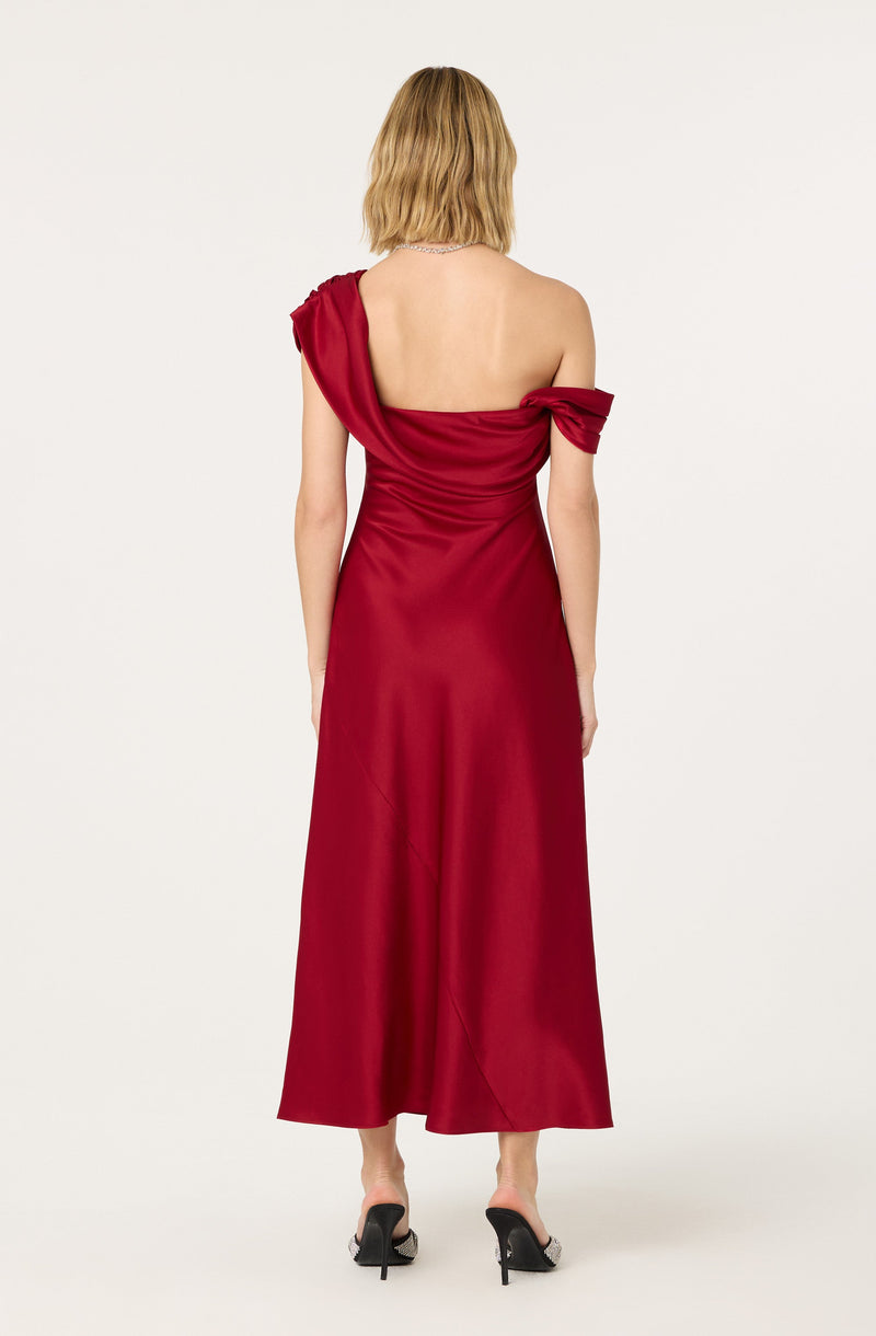 A woman is wearing a floor-length, satin gown in a deep red color. The dress features an asymmetrical design with one shoulder strap on the left side, while the right side has an off-the-shoulder sleeve that is tied in a bow. The fabric appears smooth and shiny. She is also wearing black high-heeled shoes with open toes.