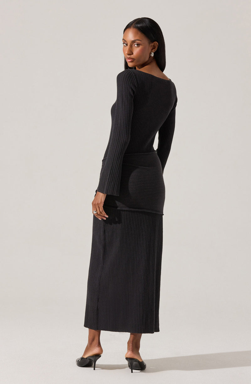 A woman is wearing a long, black, ribbed-knit dress with a fitted silhouette. The dress has a high neckline and long sleeves. She is also wearing black high-heeled shoes and earrings.