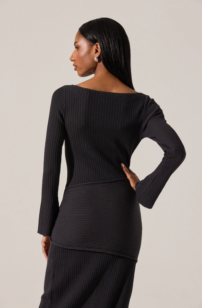 A woman is wearing a black, long-sleeved, ribbed knit top and matching ribbed knit skirt. The top has a wide neckline and the outfit appears fitted, emphasizing a sleek silhouette. She accessorizes with large, round earrings and has long, straight black hair.