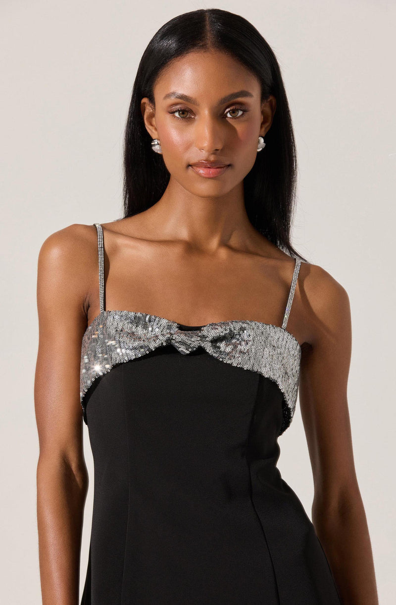 A woman is wearing a black dress with thin, sparkly straps. The upper part of the dress features a band of silver sequins and beads, emphasizing a decorative detail across the chest. The dress has a fitted silhouette, and the woman is accessorized with large pearl earrings.
