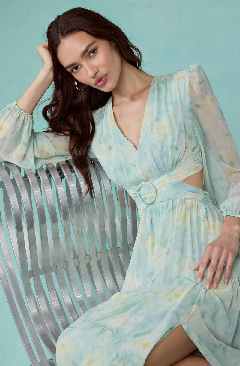 A young woman with long, dark brown hair is seated on a curved, slatted metallic bench. She is wearing a flowy, pastel-colored dress with a tie-dye pattern in soft shades of blue, yellow, and green. The dress has long, semi-sheer sleeves, a V-neckline, and a circular buckle detail at the waist, creating a cut-out effect on the sides.