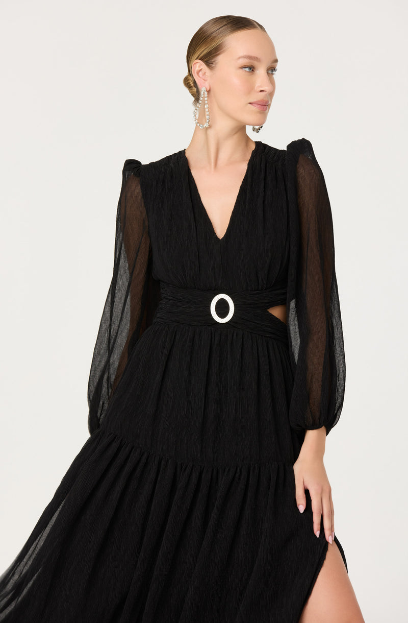 A woman is wearing a black dress with a deep V-neckline and long, sheer puffed sleeves. The dress has a wide waistband with a prominent silver oval-shaped buckle and features cutouts at the sides. The skirt of the dress is long and flowing, with pleats and a slit on one side. She accessorizes with large, looped earrings made of pearls.