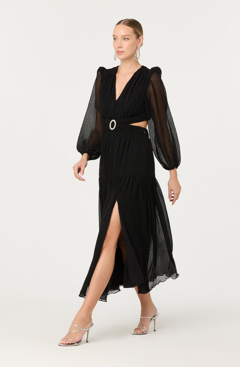 A woman is wearing a black dress with sheer, puffed long sleeves and a deep V-neckline. The dress features a fitted waist with a ring detail and a high slit that reveals her leg. She is accessorized with large hoop earrings and is wearing strappy silver high-heeled sandals.