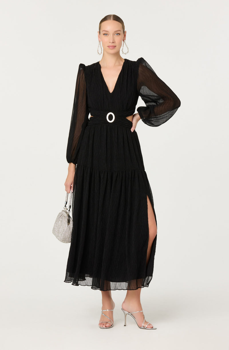 A woman is wearing a long black dress with sheer, puffed long sleeves and a V-neckline. The dress has a high slit on one side and a circular belt detail at the waist. She is accessorized with large hoop earrings, a small silver handbag, and high-heeled sandals.