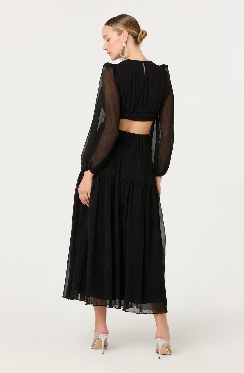 A woman is wearing a black two-piece outfit consisting of a cropped top with long, sheer puffed sleeves and a high-waisted, ankle-length skirt. The top has a loose, gathered design at the bust and a button closure at the back. The skirt is flowing with a tiered, ruffled style. She accessorizes with large, dangling, pearl earrings and white high-heeled shoes. Her hair is styled in a low, sleek bun.