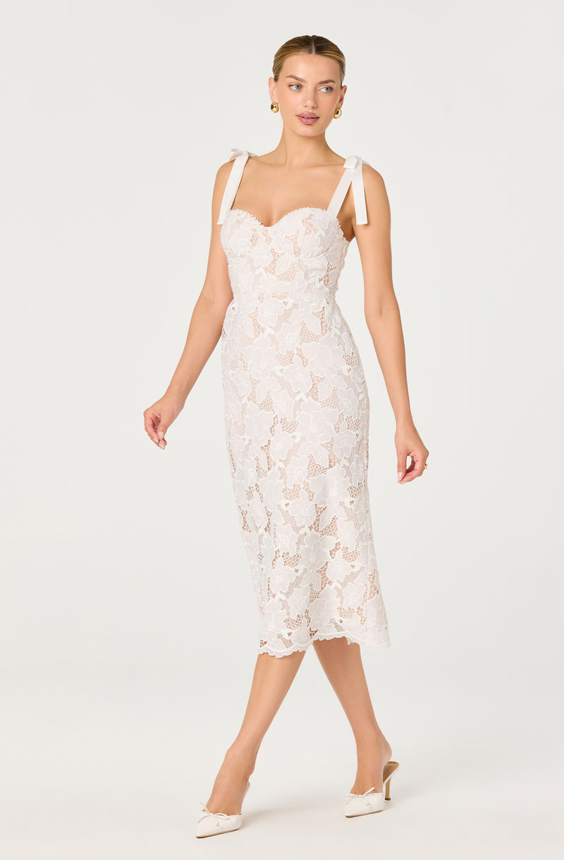 Woman wearing a white lace midi dress with bow shoulder straps and cream-colored heels.