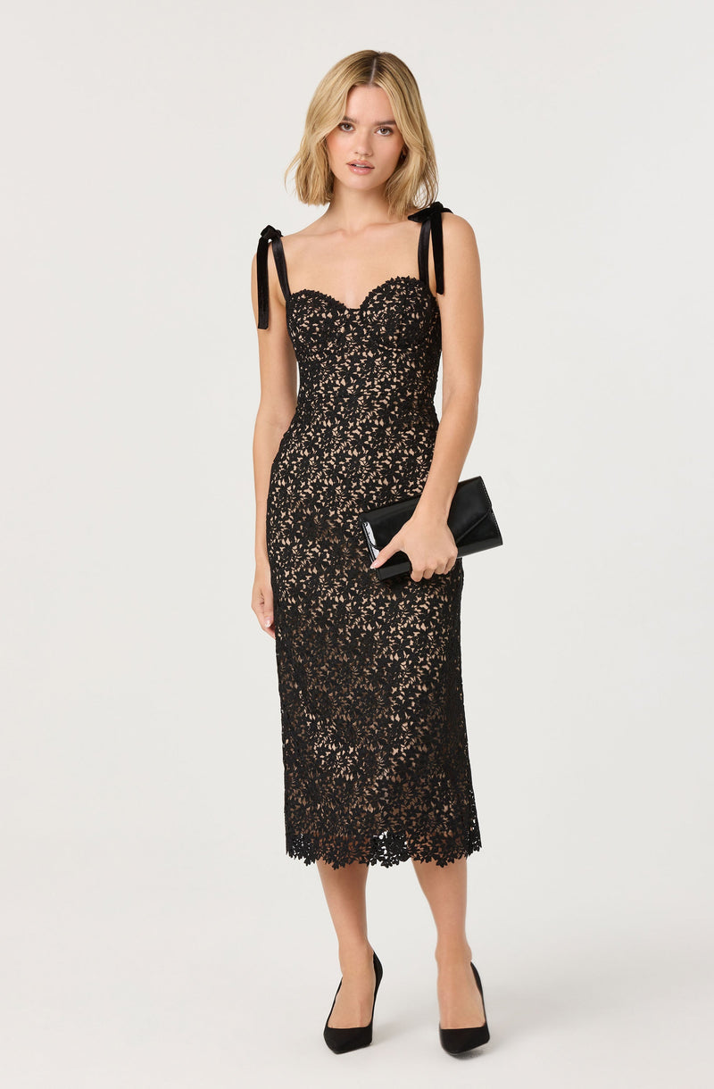 A woman is wearing a black lace midi dress with a fitted silhouette. The dress has a sweetheart neckline and is accented with black fabric shoulder ties that form bows on each shoulder. She is holding a black clutch and wearing black high-heeled shoes.