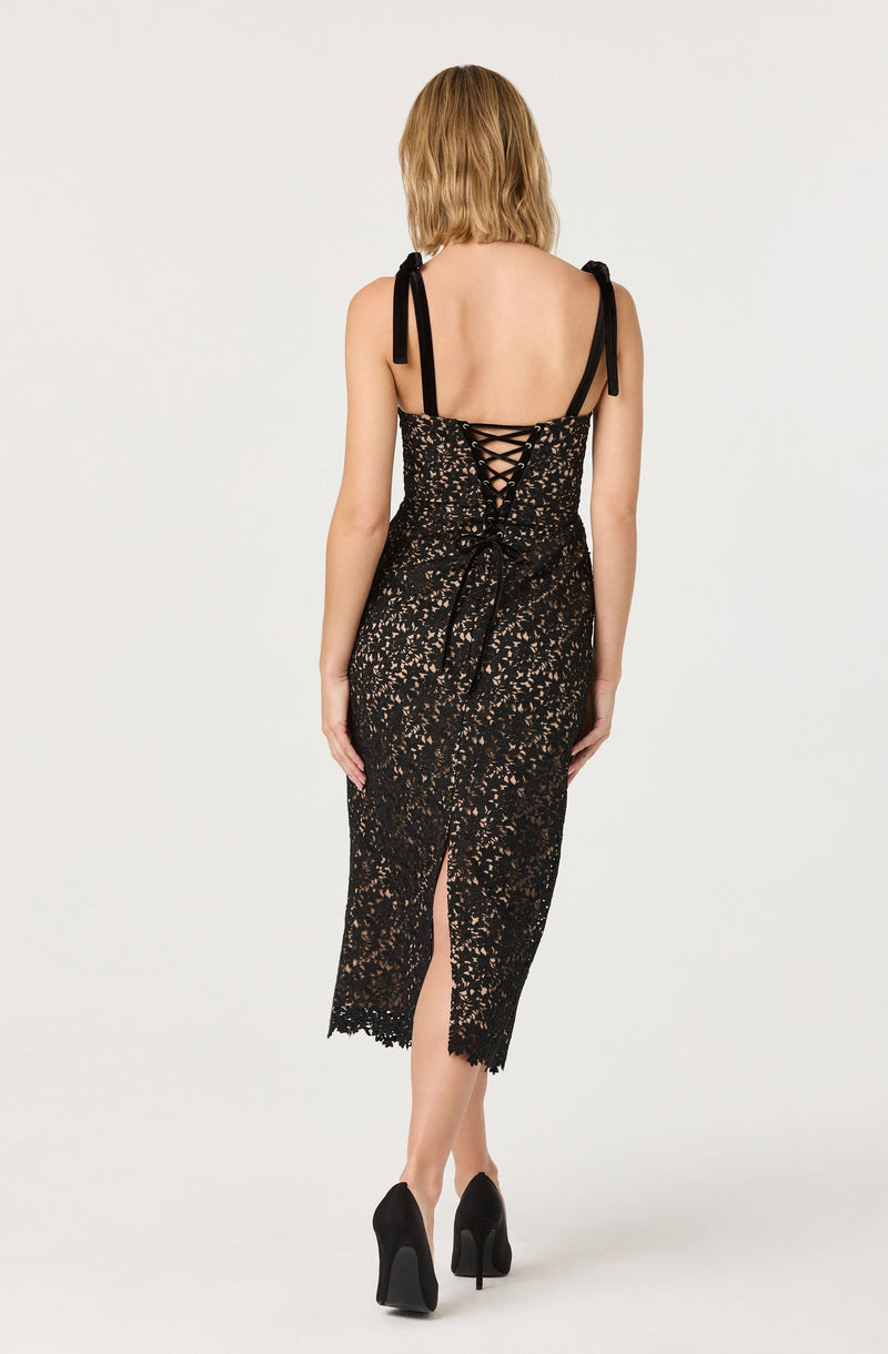 The dress is a black, form-fitting, sleeveless garment with thin shoulder straps tied in bows. It features a lace-up back detail with crisscrossing black laces and has a slit at the back hem. The fabric has a floral lace overlay with a nude or beige lining underneath, creating a see-through effect on the lower part of the dress. The model is wearing black high-heeled shoes.