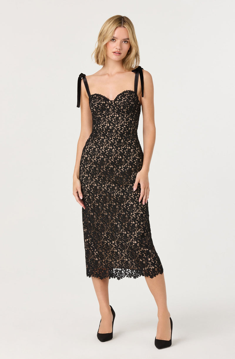 The dress is a black lace midi-length garment with a fitted silhouette. It features a sweetheart neckline and thin shoulder straps tied into bows at the shoulders. The lace overlay has a floral pattern, creating a semi-transparent effect over a nude lining. The hemline reaches below the knees with a scalloped lace edge. The model is wearing black high heels with the dress.