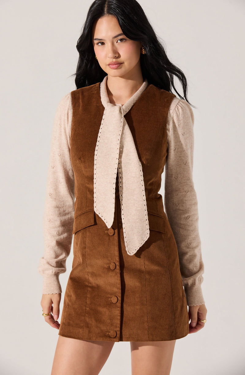 A woman is wearing a brown corduroy sleeveless dress with large buttons down the front. Underneath, she has on a beige long-sleeve sweater featuring a subtle heart pattern. She also has a matching beige scarf tied loosely around her neck.