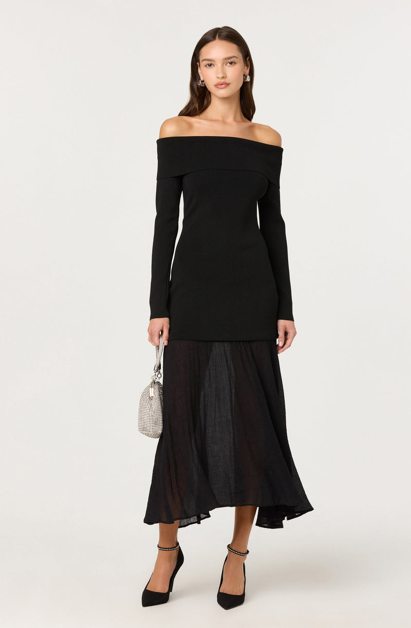 A woman is wearing a black off-the-shoulder dress with long sleeves and a fitted bodice that extends to mid-thigh. She is also wearing a black, semi-sheer, ankle-length skirt underneath. She accessorizes with a small, textured silver handbag, hoop earrings, and black high-heeled shoes with ankle straps.