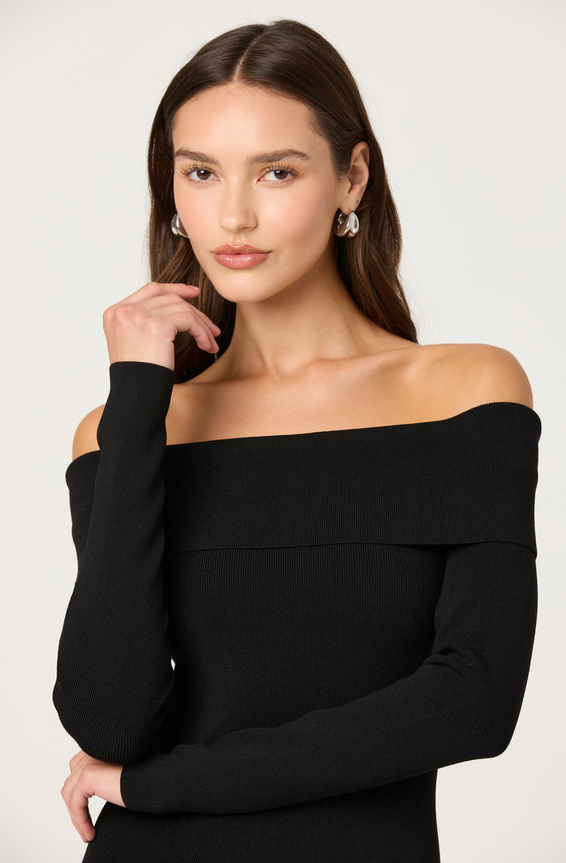 A woman wearing an off-the-shoulder black top with long sleeves. She has on large, shiny hoop earrings and has her hand resting near her face.