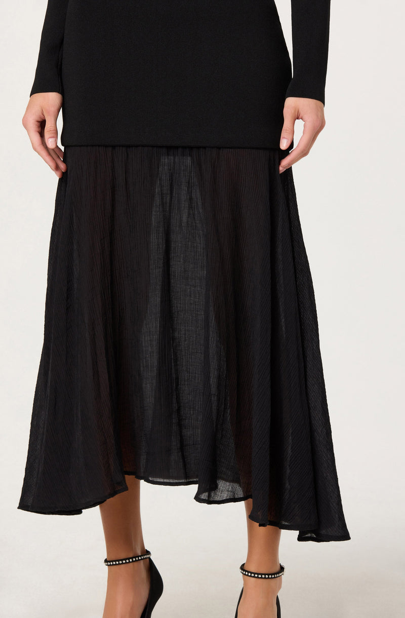 A woman is wearing a long black pleated skirt made of lightweight, semi-sheer fabric. She pairs it with a black long-sleeve top that extends slightly below the waist. She is also wearing black high-heeled shoes with ankle straps decorated with small pearls.