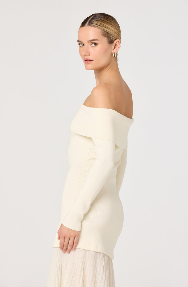 Off-shoulder cream dress with a fold-over neckline and long sleeves, paired with a light-colored skirt.