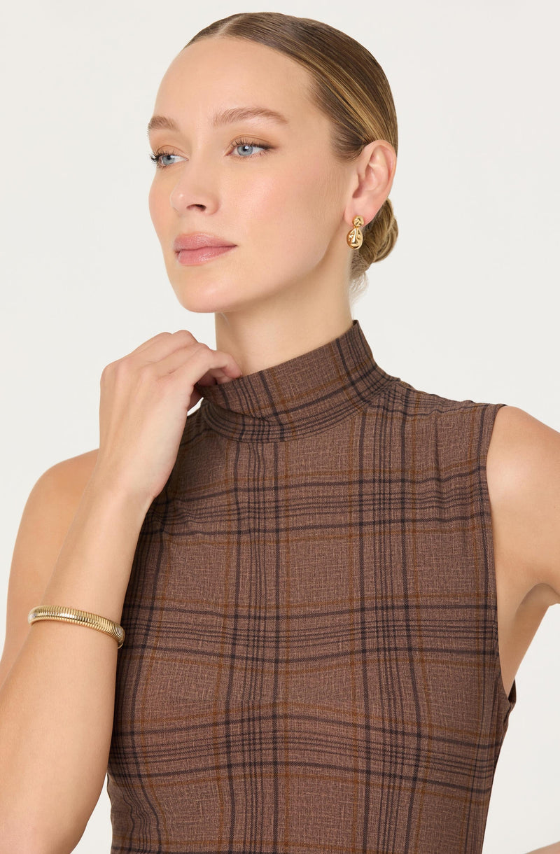 A woman is wearing a sleeveless, high-neck top with a brown and orange plaid pattern. She has gold earrings and a gold bracelet on her right wrist. Her hair is pulled back in a neat bun.
