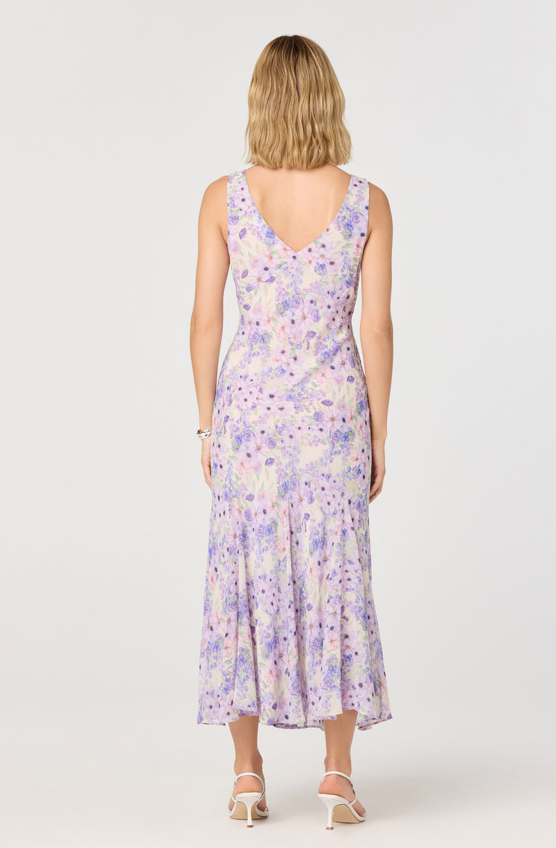 The dress is sleeveless with wide shoulder straps and a deep V neckline at the back. It features a pastel-colored floral and abstract pattern with shades of purple, lavender, pink, and cream. The fabric appears lightweight with a flowy silhouette, fitted at the waist, and flares out towards the hem, which reaches mid-calf length.