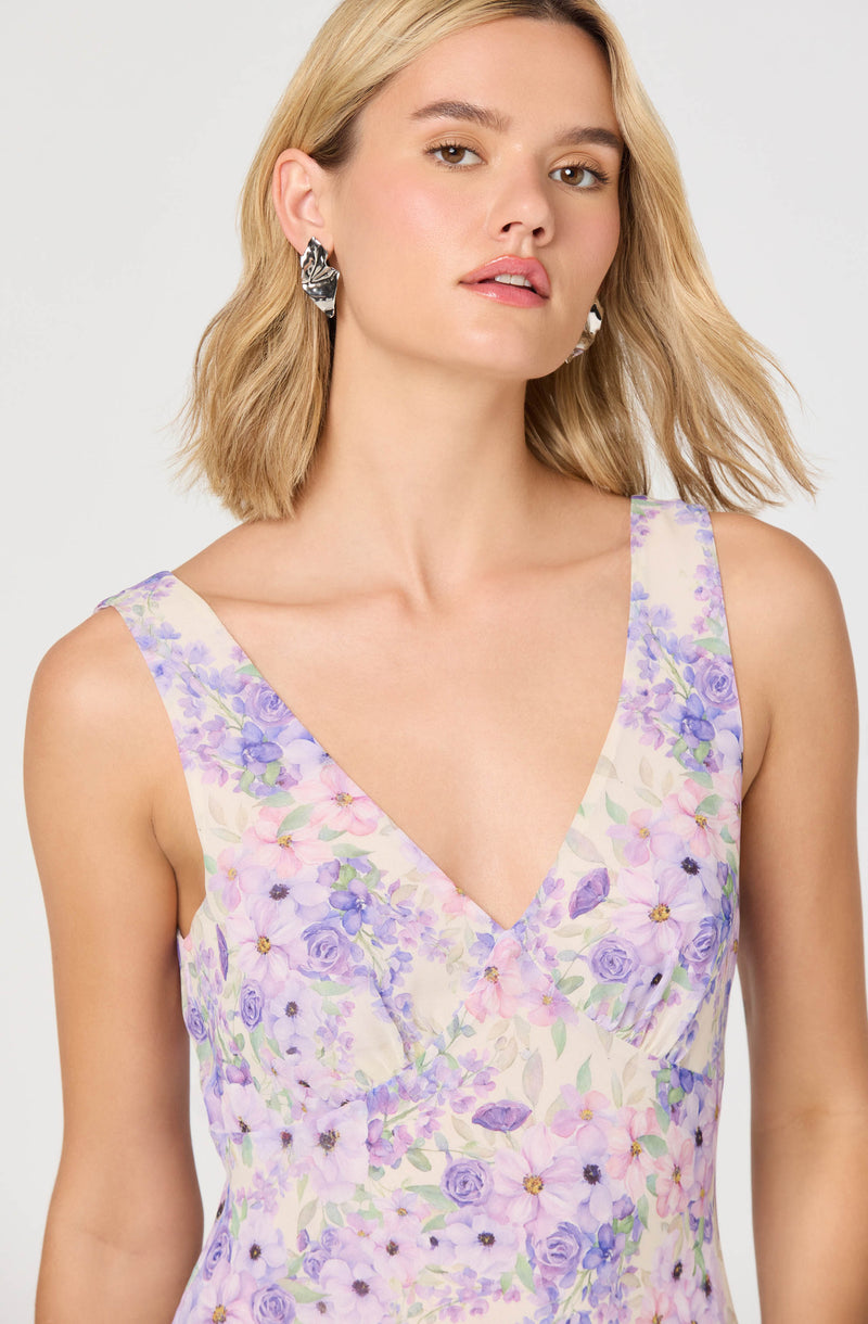 A woman is wearing a sleeveless dress with a V-neckline. The dress features a floral pattern with light purple, lavender, pink, and green colors on a light background. She has short, wavy blonde hair and is accessorized with large, abstract black and white earrings.