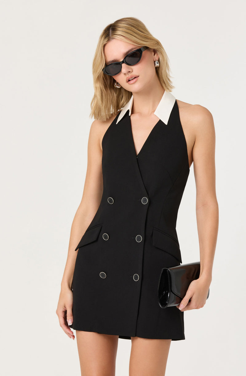 A woman is wearing a sleeveless black dress with a V-neckline and double-breasted button details. The dress features a white collar, adding a contrast to the black fabric. She accessorizes with black sunglasses, silver earrings, and holds a glossy black clutch in her right hand.