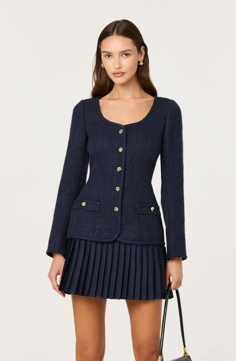 A woman is wearing a navy blue textured long-sleeve blazer with a rounded neckline. The blazer has gold buttons down the front and on the small decorative pockets at the hips. She is also wearing a matching navy blue pleated skirt.