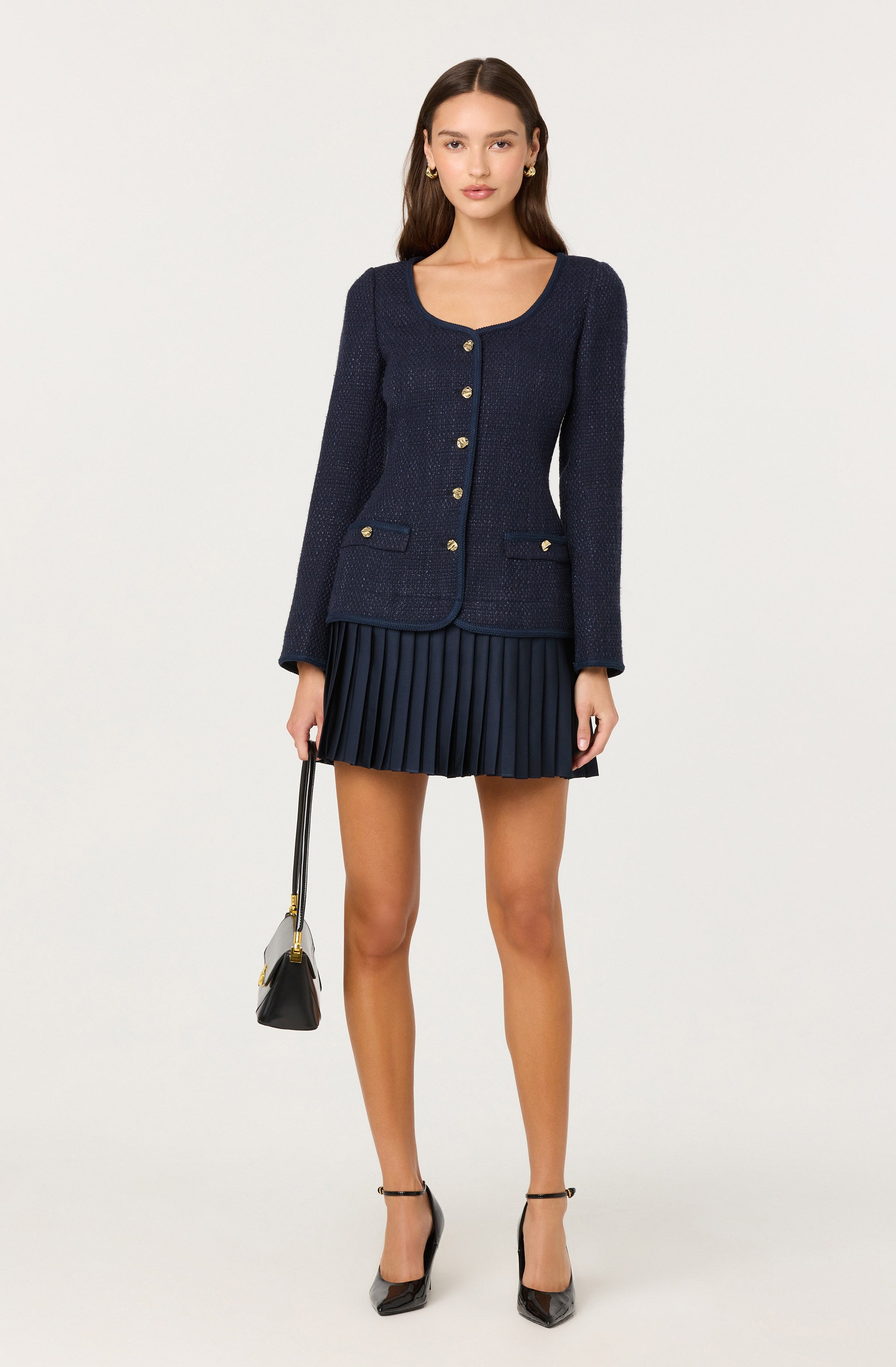 A woman is wearing a fitted navy blue textured blazer with long sleeves, gold buttons down the front, and decorative pockets on the chest. She pairs it with a matching navy blue pleated mini skirt. She is also wearing black high-heeled shoes and holding a small black handbag with a shoulder strap.