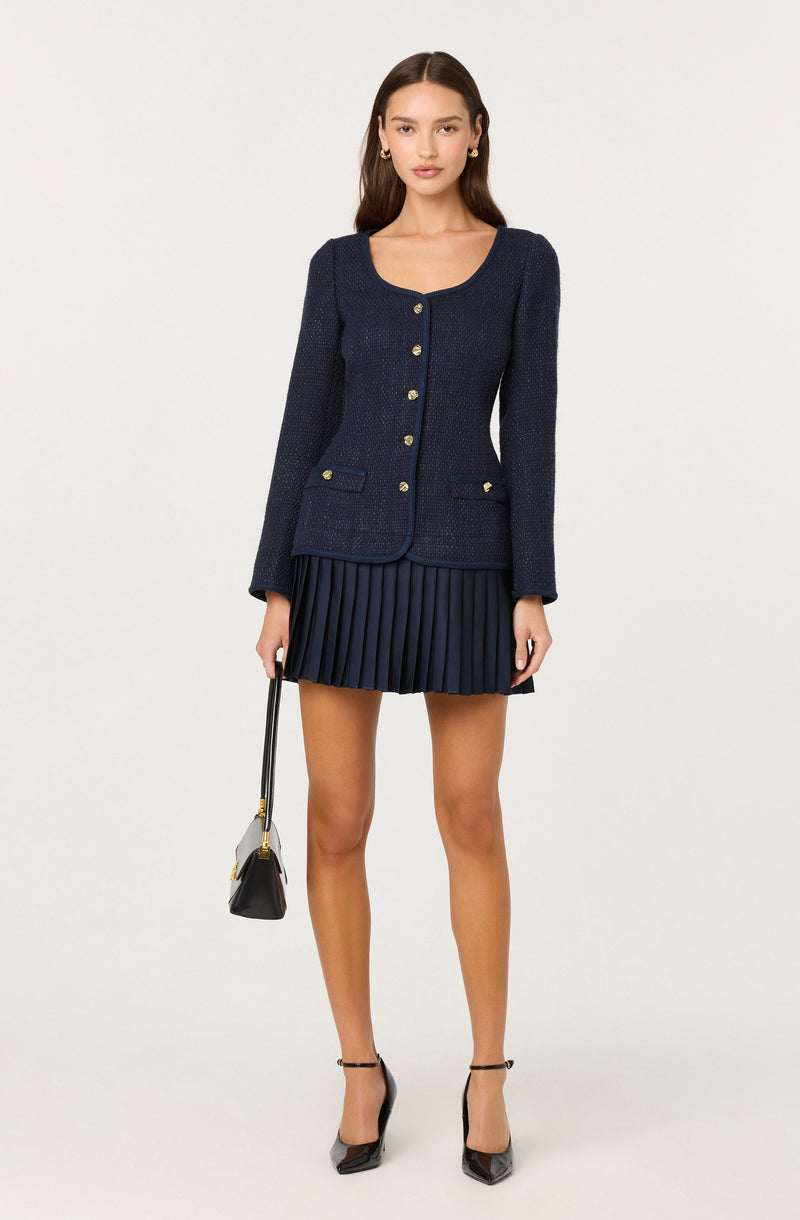 A woman is wearing a fitted navy blue textured blazer with long sleeves, gold buttons down the front, and decorative pockets on the chest. She pairs it with a matching navy blue pleated mini skirt. She is also wearing black high-heeled shoes and holding a small black handbag with a shoulder strap.