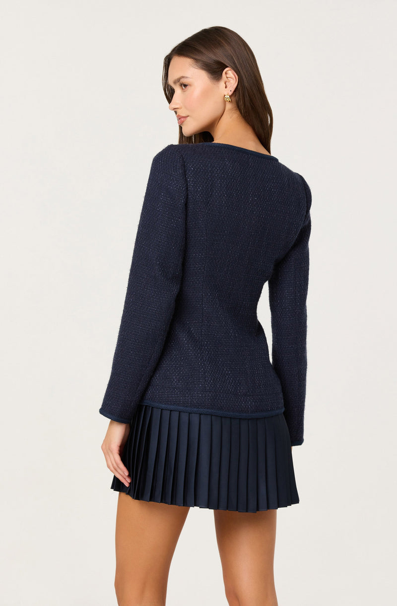 A woman is wearing a navy long-sleeved, textured blazer and a matching pleated skirt. The blazer is fitted with a simple, clean design, and the skirt features evenly spaced vertical pleats. The overall look is elegant and sophisticated.