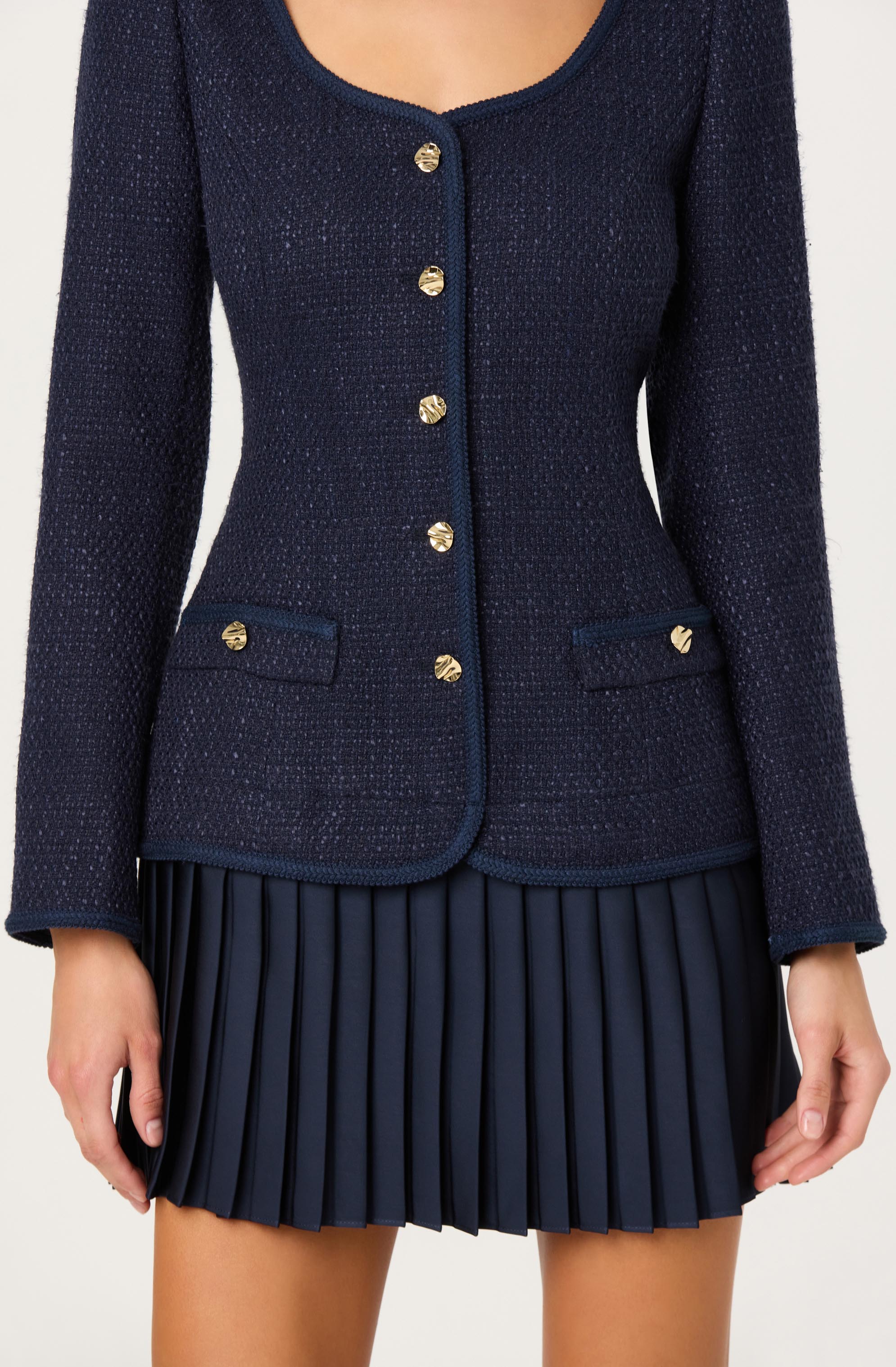 A woman is wearing a navy blue textured blazer with gold buttons down the front and on the two small pockets at the hips. The blazer has a round neckline with a slight scoop, and the edges are finished with trim. She is also wearing a matching navy blue pleated skirt with evenly spaced vertical pleats.
