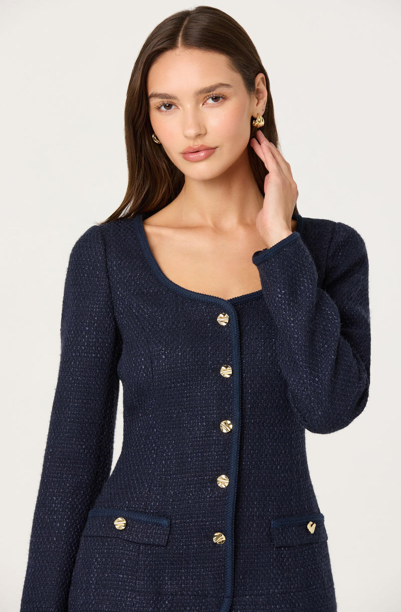 A woman is wearing a dark navy, textured, long-sleeve jacket with gold buttons down the front and on the pockets. The jacket has a rounded neckline and a fitted silhouette. She is accessorized with gold earrings.