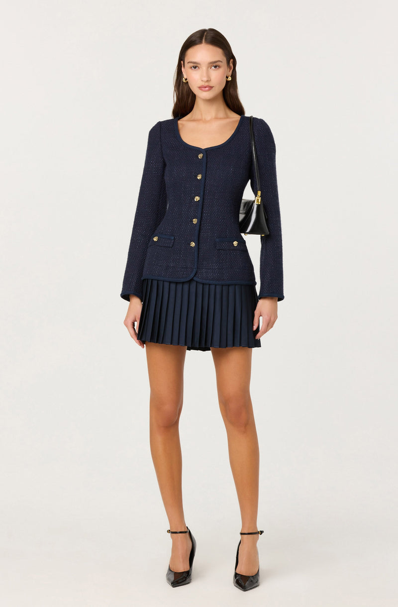 A woman is wearing a navy blue textured blazer with gold buttons down the front, on the pockets, and at the cuffs. She pairs it with a matching navy blue pleated mini skirt. She has a black shoulder bag with gold hardware, and she is wearing black high-heeled shoes with ankle straps.
