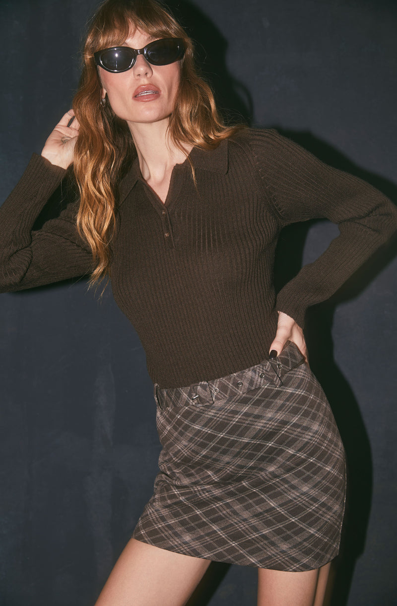 Woman wearing a brown ribbed sweater and plaid skirt against a dark background