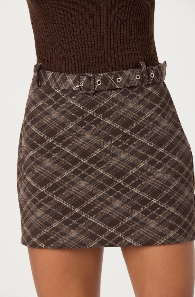 A woman is wearing a high-waisted, brown plaid skirt with a matching belt featuring metal eyelets and a tie. She pairs it with a fitted, long-sleeved, ribbed brown sweater.