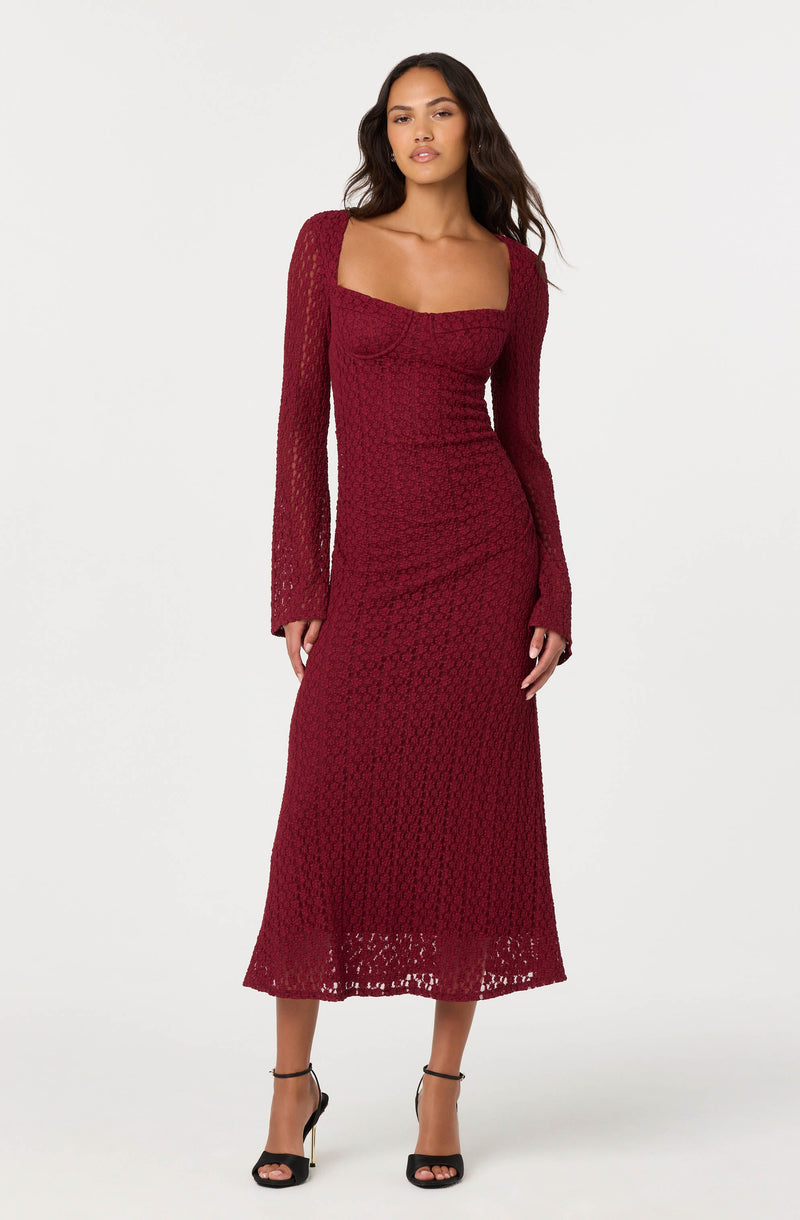 A woman is wearing a long, deep red lace dress with long sleeves. The dress features a square neckline and a fitted silhouette that extends to a mid-calf length. She is also wearing black high-heeled sandals with ankle straps.