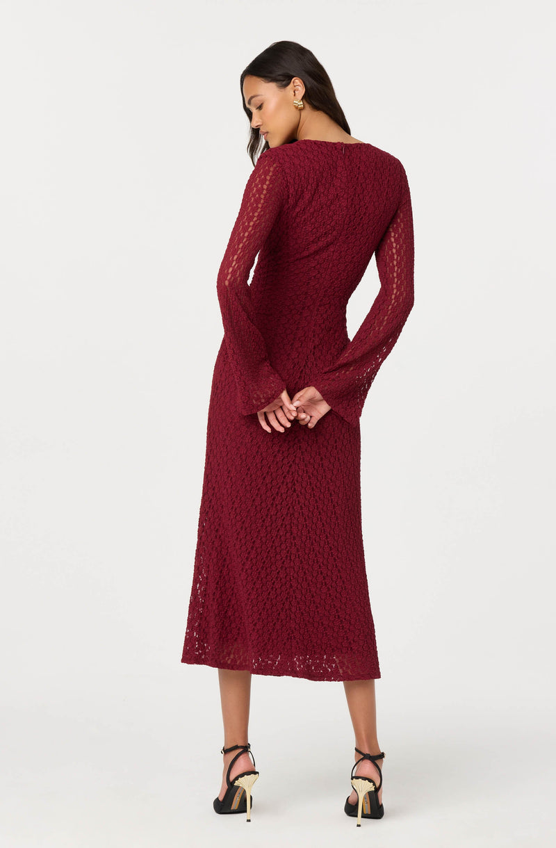 A woman is wearing a long, deep red lace dress with long, semi-sheer sleeves and a fitted silhouette. She is also wearing black high-heeled sandals with ankle straps and gold accents.