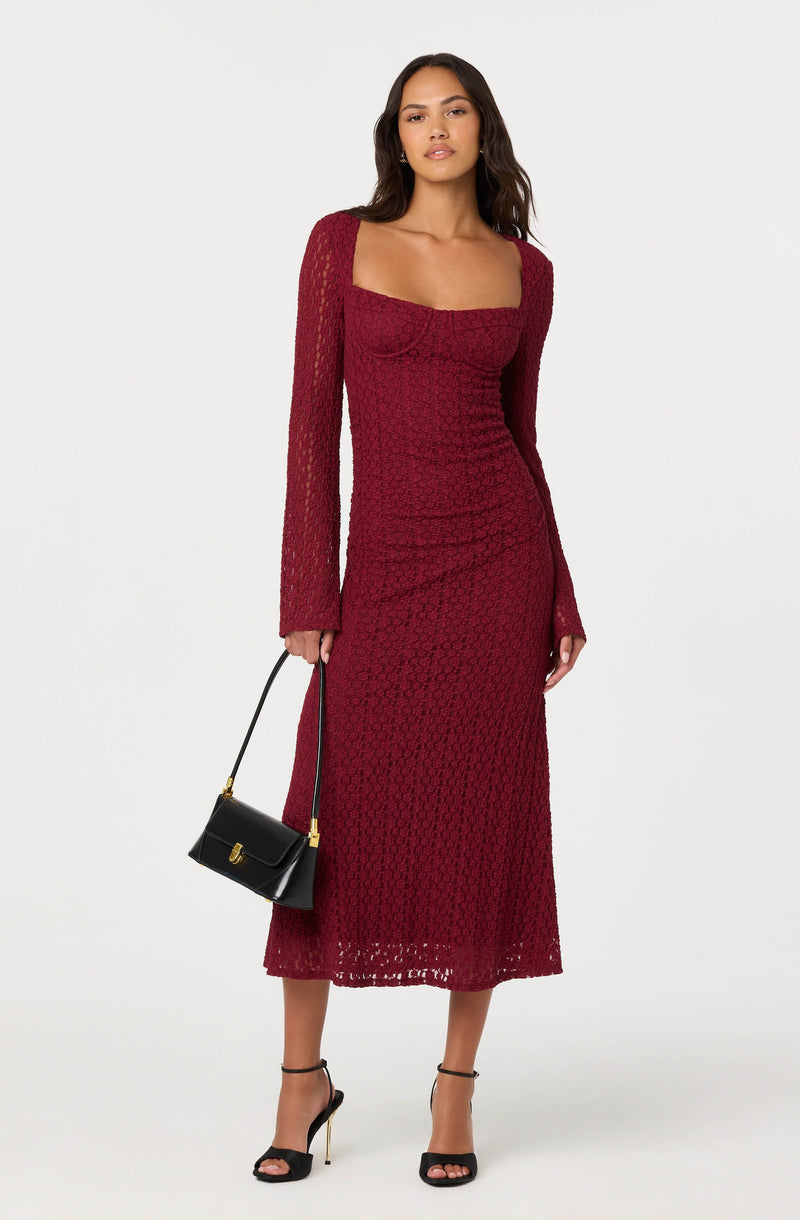A woman wearing a long, red, textured lace dress with long sleeves and a square neckline. She is holding a small black handbag with a gold clasp and is wearing black high-heeled sandals with ankle straps.