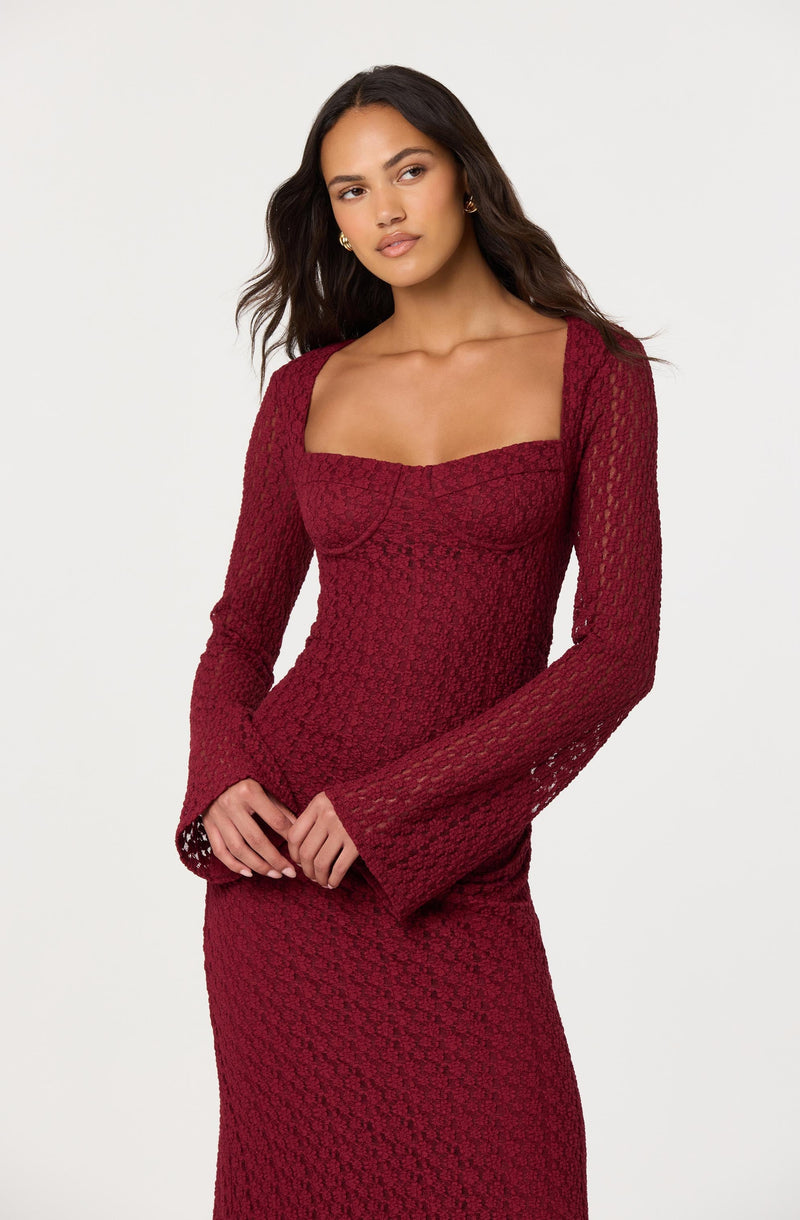 A woman is wearing a fitted, long-sleeved dress in a deep red color. The dress has a square neckline and features a textured, lace-like pattern throughout. The sleeves are slightly flared at the cuffs.