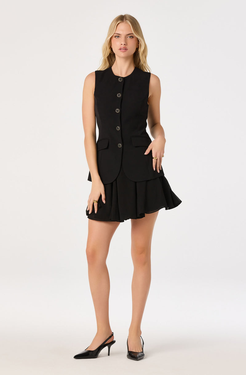 Woman wearing a sleeveless black blazer with decorative buttons and a matching ruffled black skirt.