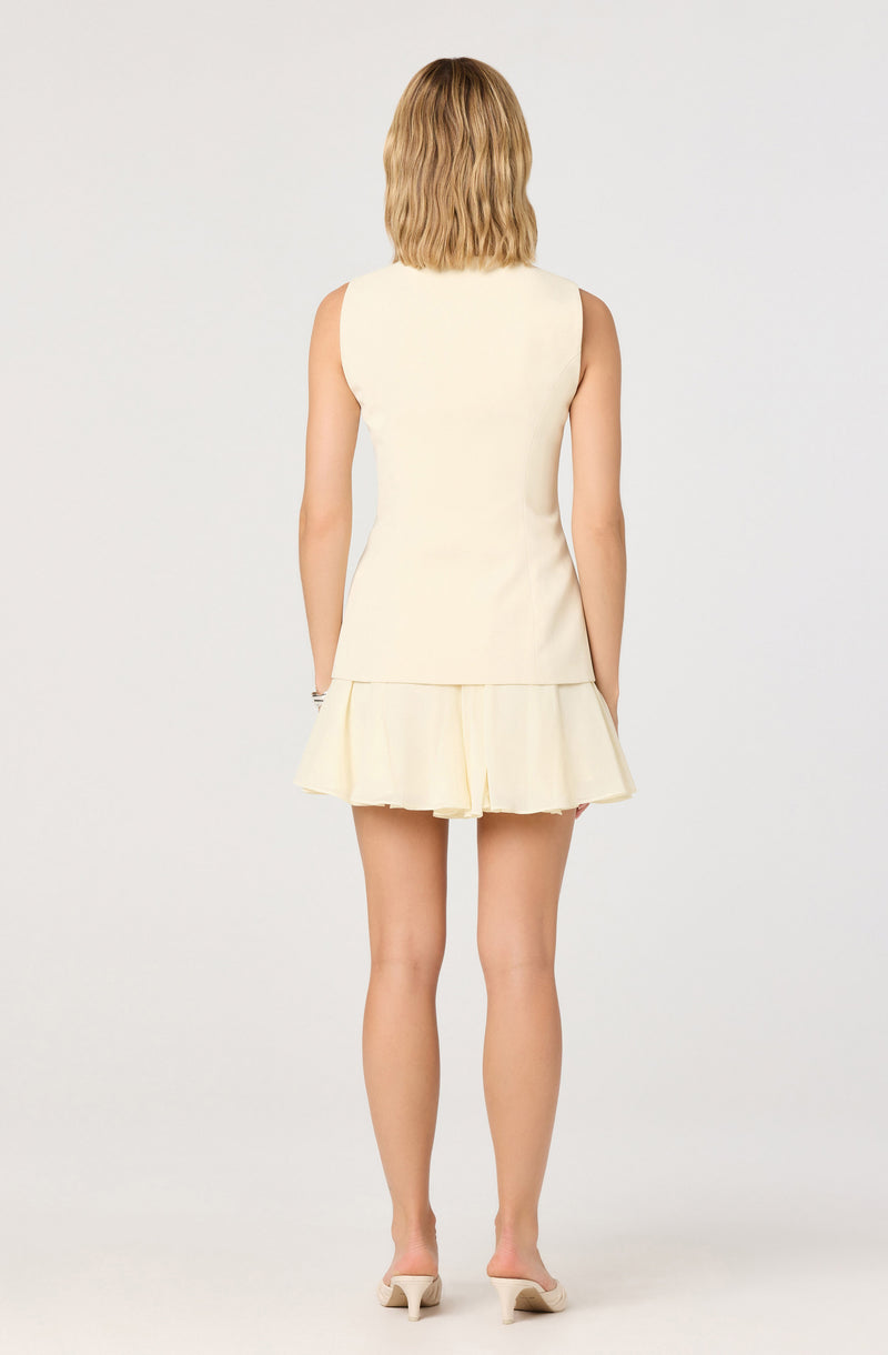 The woman is wearing a sleeveless, cream-colored top with a fitted bodice and a matching short skirt with a ruffled, layered hem. She has blonde, shoulder-length hair and is standing barefoot against a plain light background.