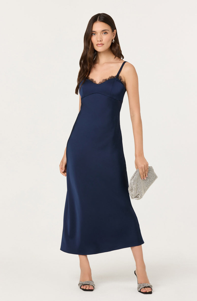 A woman is wearing a navy blue slip dress with thin spaghetti straps and lace trim along the neckline. She is holding a silver, textured clutch bag and wearing high-heeled sandals with snake print details.