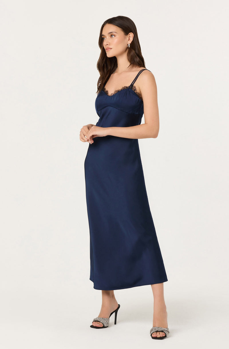 A woman is wearing a navy blue satin slip dress with spaghetti straps and lace trim along the neckline. She is also wearing high-heeled sandals with an embellished strap.