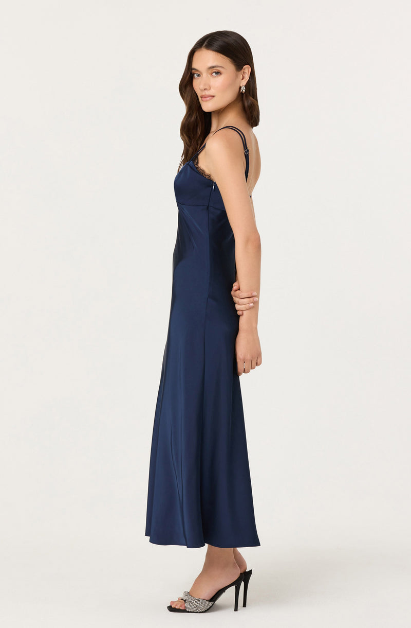 A woman is wearing a navy blue satin slip dress with thin, adjustable spaghetti straps and lace trim along the top edge. She is also wearing black high-heeled sandals with an open toe and embellished straps.
