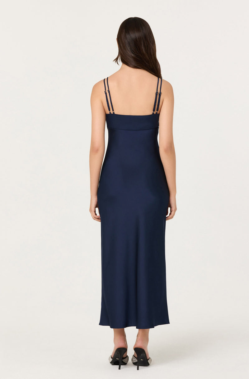 A woman is wearing a navy blue, sleeveless, full-length dress with thin, double straps on her shoulders. The dress is fitted at the top and flows straight down to the hem. She is also wearing black high-heeled shoes.