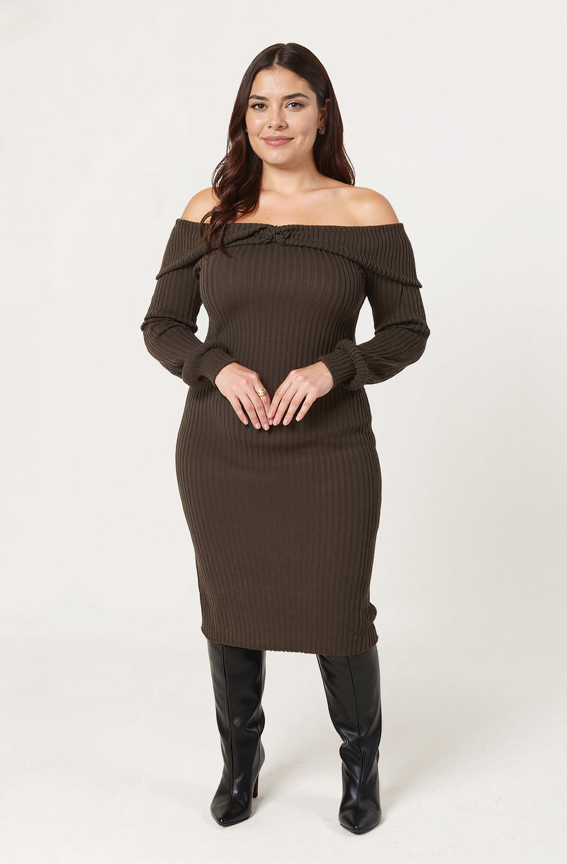 A woman is wearing an off-the-shoulder, long-sleeved, ribbed sweater dress in a dark olive green color. She is also wearing tall, black leather boots.