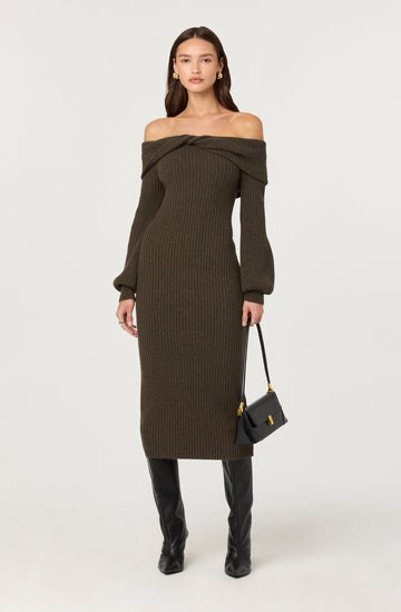 A woman is wearing an off-the-shoulder, long-sleeved knit dress in a dark olive green color. The dress is form-fitting and extends to mid-calf. She is holding a small black handbag with gold accents and is wearing black knee-high boots.
