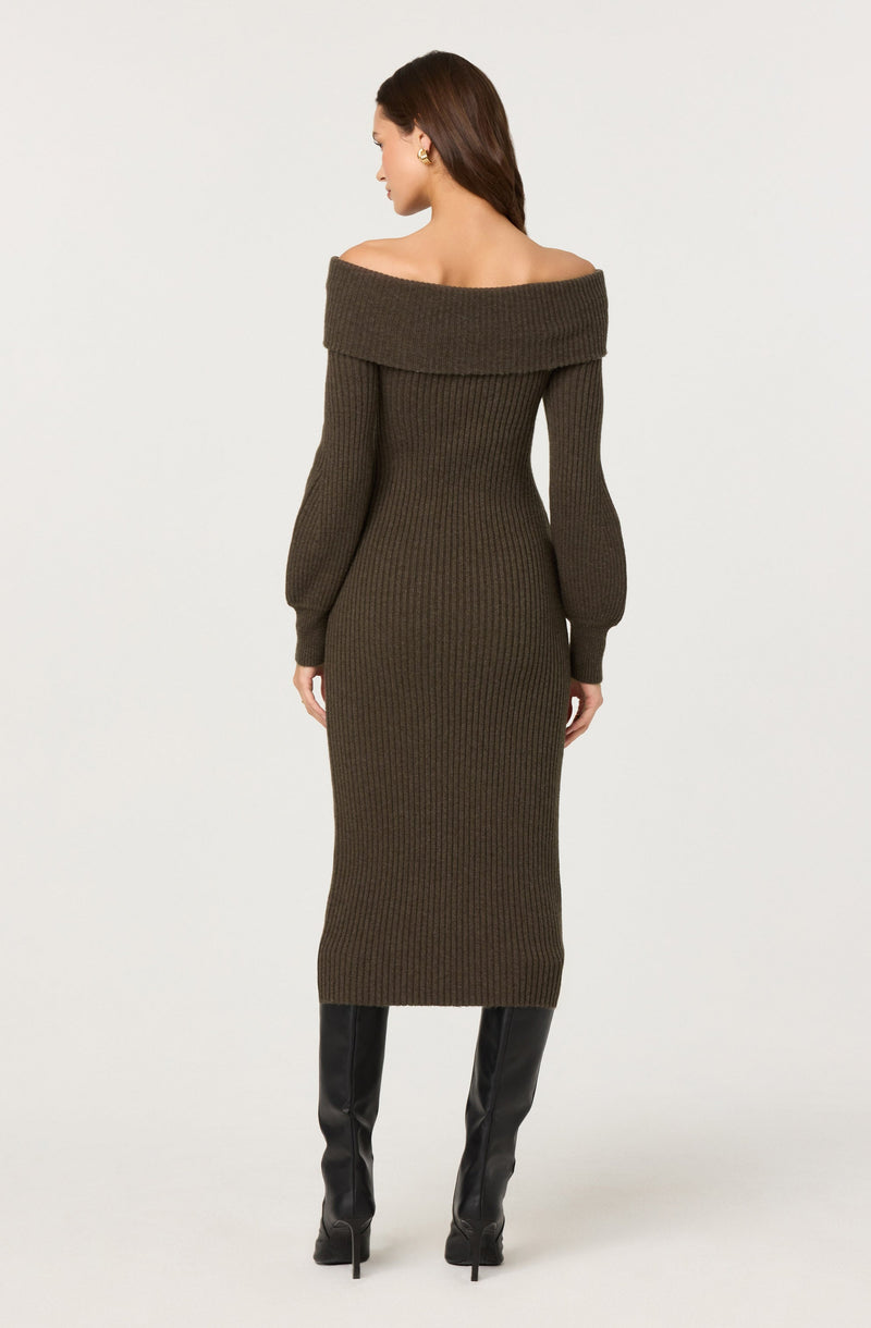 A woman is wearing a long, dark brown, ribbed knit sweater dress with an off-the-shoulder neckline that folds over the shoulders. The dress has long sleeves with fitted cuffs at the wrists. She is also wearing tall black boots that reach just below the knees.
