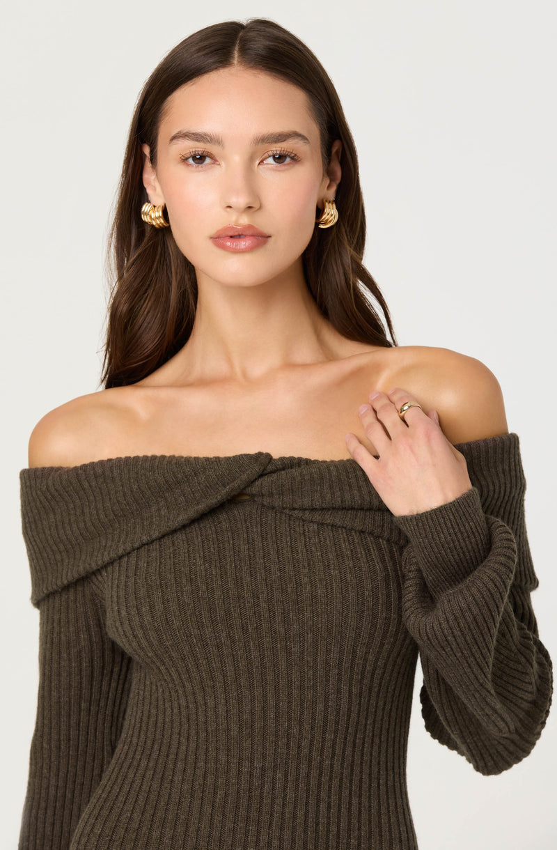 A young woman with long, wavy brown hair is wearing an off-the-shoulder, ribbed, olive-green sweater that fits snugly. She has gold hoop earrings and a gold ring on her left hand. Her expression is neutral, and she is looking directly at the camera against a plain white background.