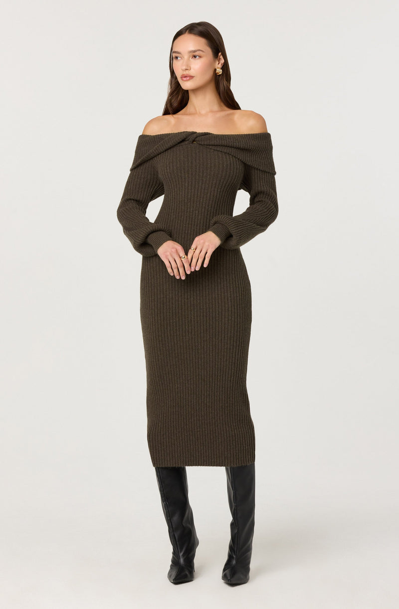 A woman is wearing a dark olive green, ribbed, off-the-shoulder knit dress that extends to mid-calf length. The dress features a wide neckline that drapes off her shoulders and long sleeves. She has on black knee-high boots and accessorizes with gold earrings and rings.