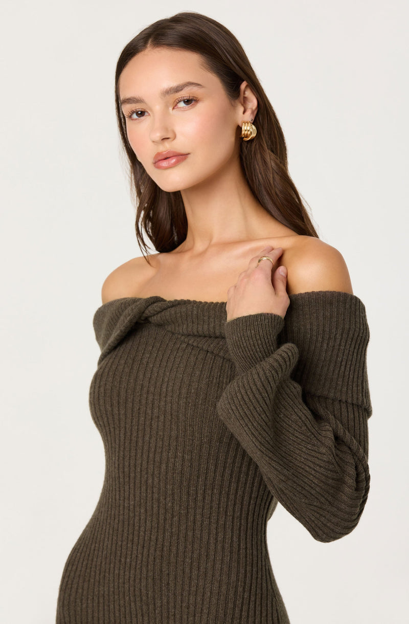 A young woman with long brown hair is wearing a dark olive green, off-the-shoulder, ribbed knit sweater with long, loose sleeves. She is accessorized with gold hoop earrings and a gold ring on her right hand.