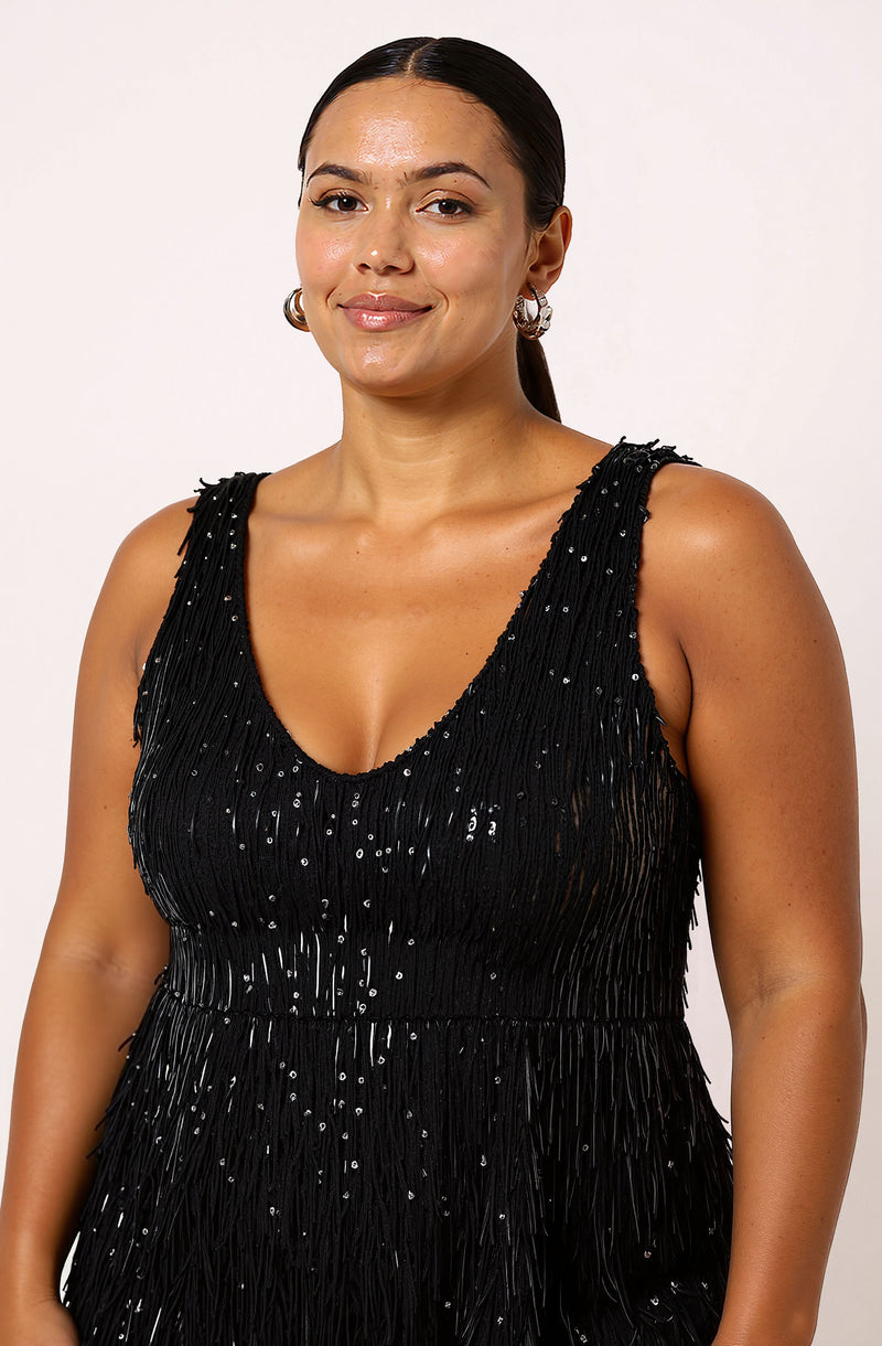 A woman is wearing a sleeveless black dress made of fabric adorned with shiny, vertical fringe and small, scattered sequins. The dress has a deep V neckline and a fitted bodice. She accessorizes with large hoop earrings.