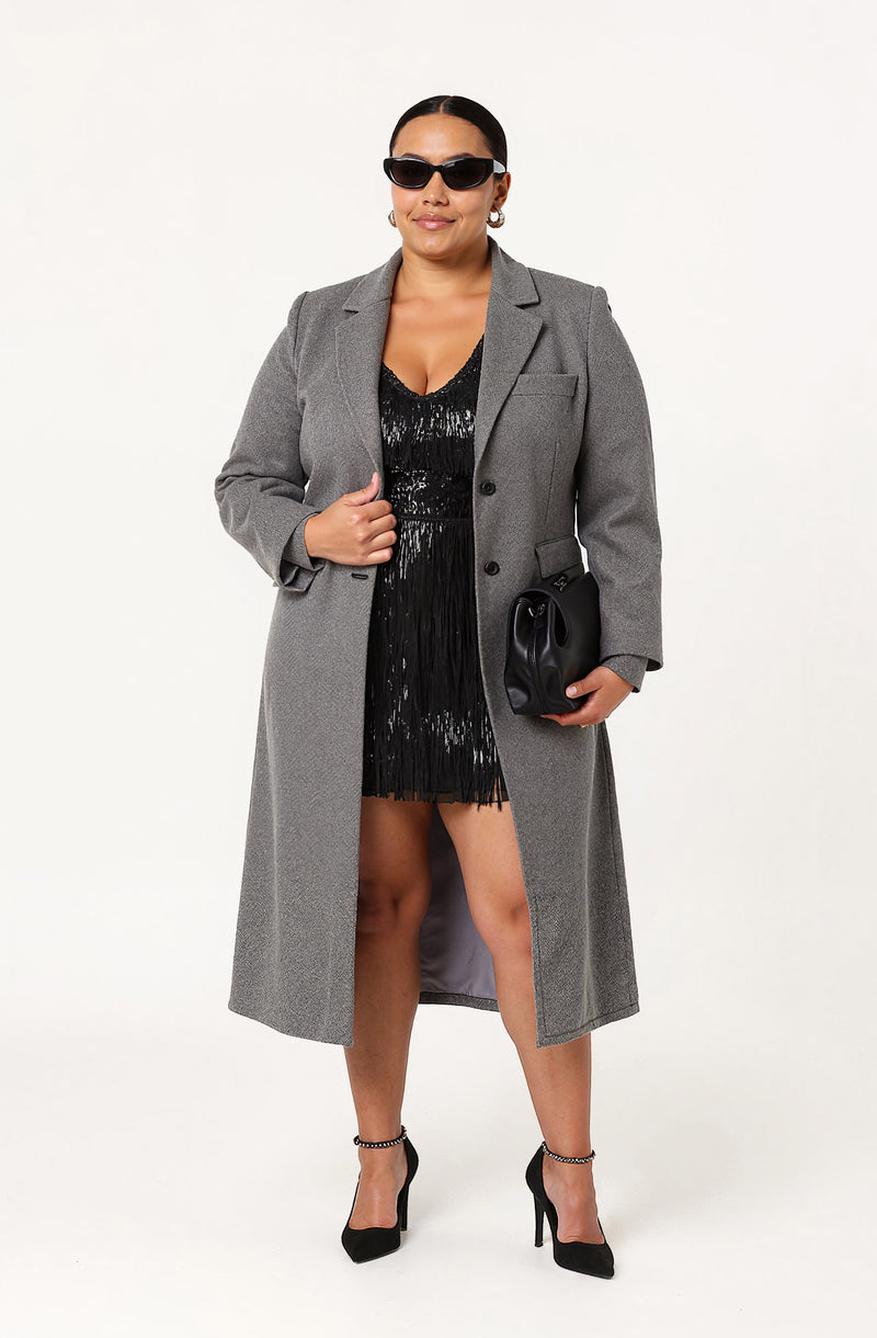 A woman is wearing a long gray blazer with a tailored fit and lapels, over a black sequined dress with a deep neckline and ruching details. She pairs the outfit with black high-heeled shoes that have ankle straps, black sunglasses, and hoop earrings, and holds a small black clutch purse.