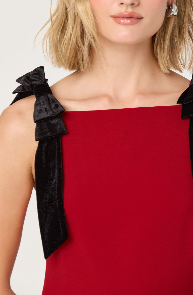 A woman is wearing a sleeveless red dress with black velvet shoulder straps tied into bows.
